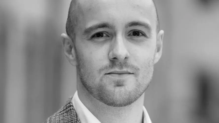 Hloedver has shaved hair and is wearing a checked blazer and white shirt