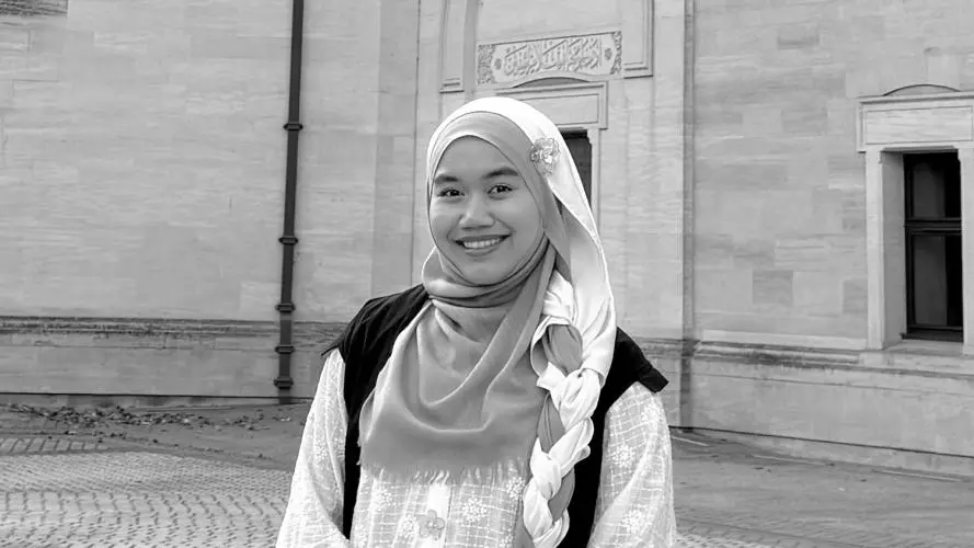 Anis Amran is standing outside in a large courtyard, wearing a subfusc gown over her clothing