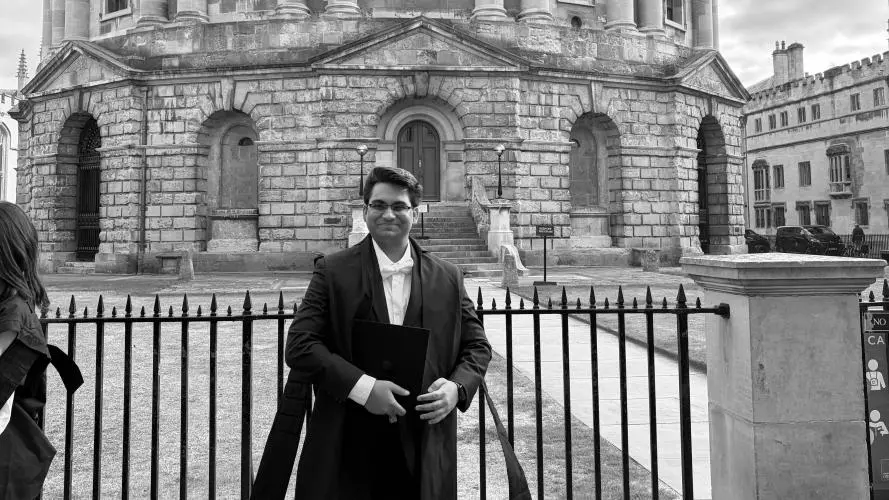 Muhammad is standing in front of the Radcliffe Camera in Oxford, wearing subfusc clothing