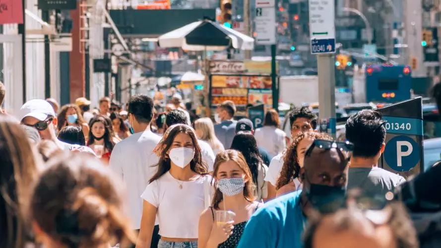 People walk along a crowded street, wearing masks
