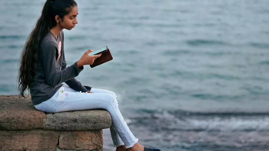 Image of a young woman sitting near the sea using a mobile phone