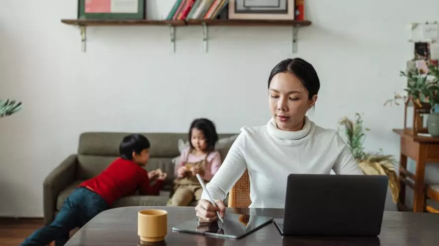 Young mother works remotely while children play on sofa behind