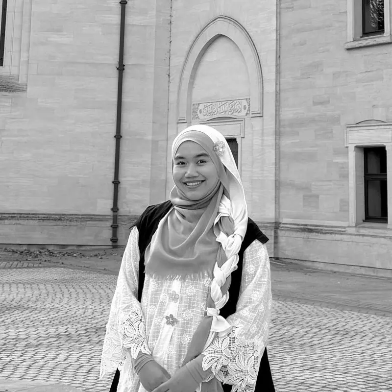 Anis Amran is standing outside in a large courtyard, wearing a subfusc gown over her clothing