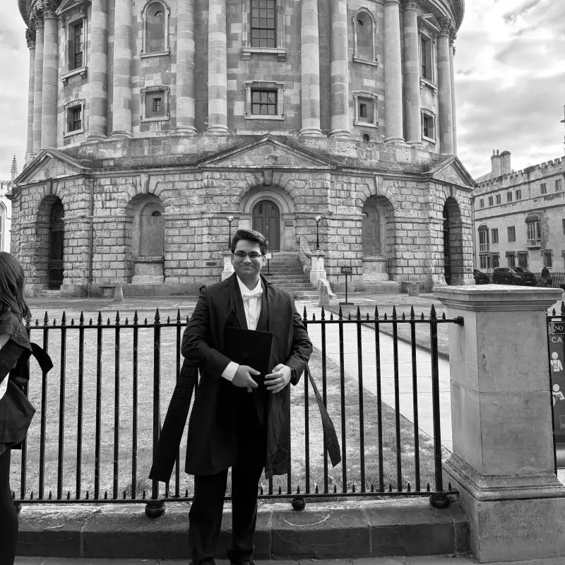 Muhammad is standing in front of the Radcliffe Camera in Oxford, wearing subfusc clothing
