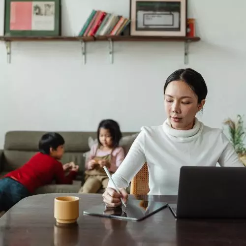 Young mother works remotely while children play on sofa behind